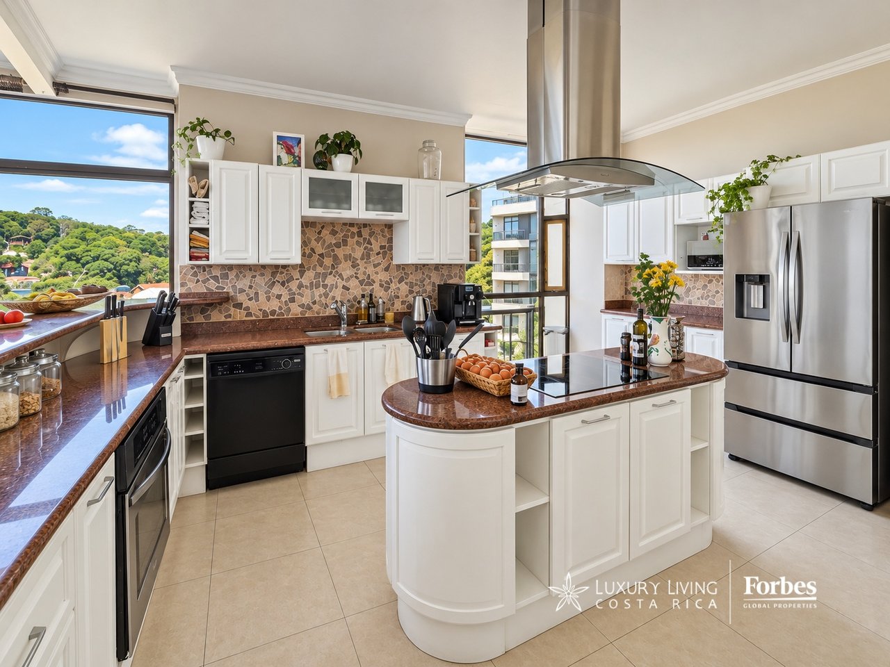 Penthouse kitchen with a rounded cooktop island, patterned stone backsplash, and views of wooded hills.