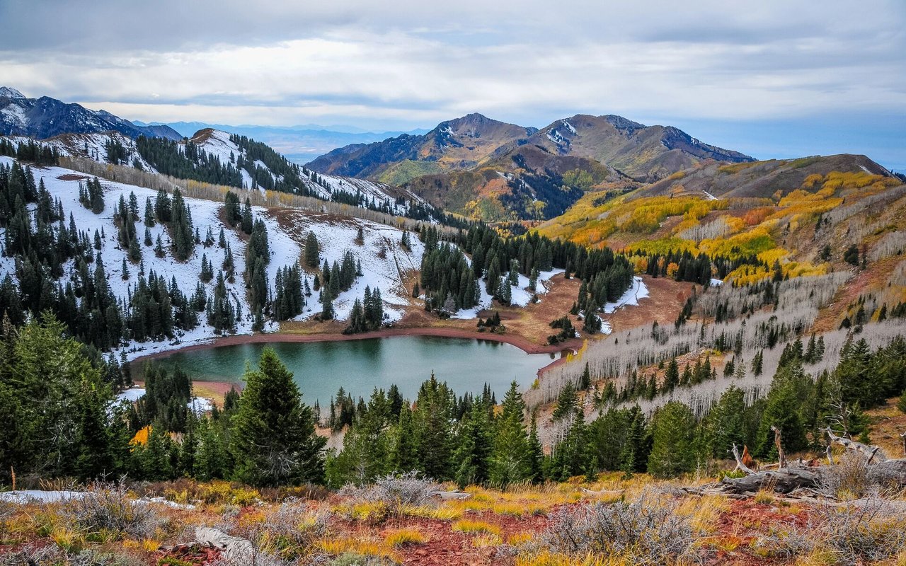 Alpine lake surrounded by evergreen forest, autumn-colored mountains, and patches of snow.
