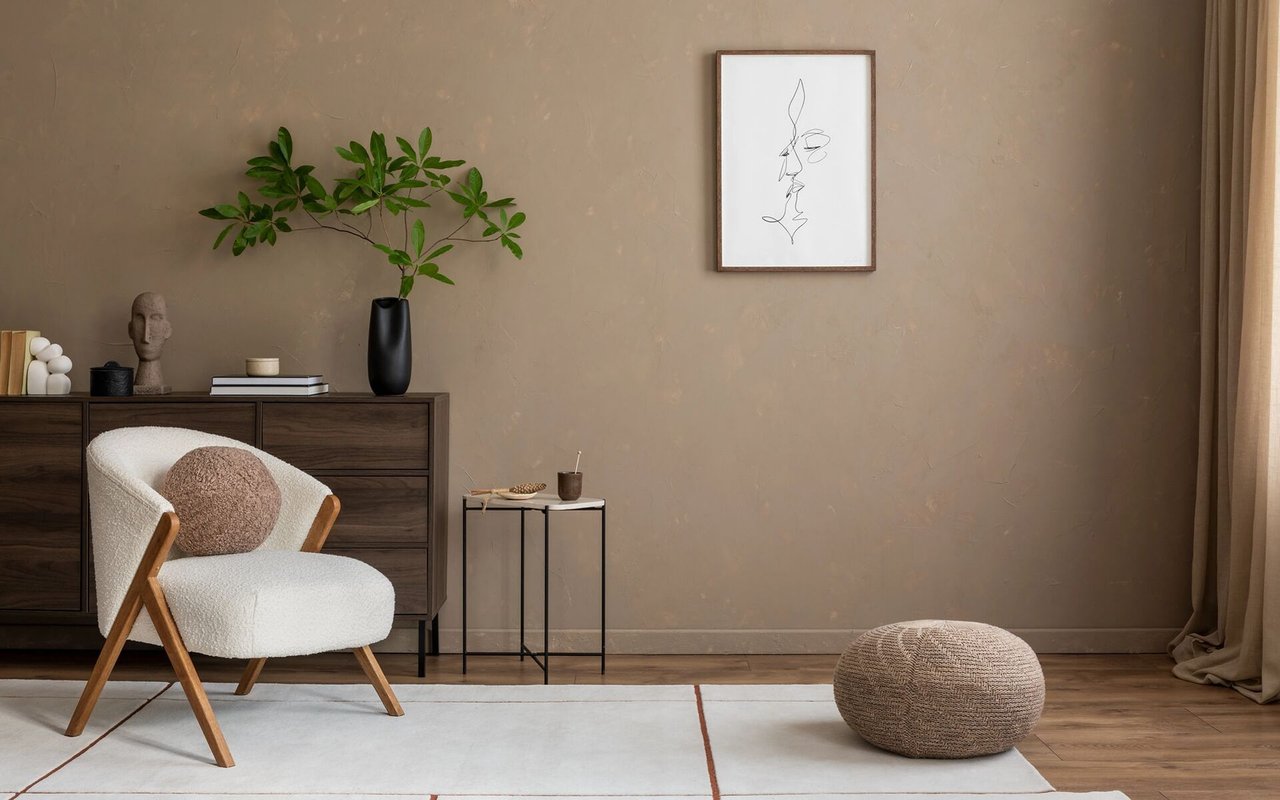 Furnished living room with a white lounge chair, dark wood dresser, leafy branches, and woven pouf.