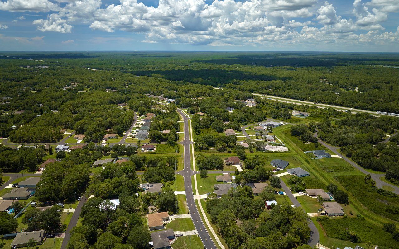Aerial view of a residential neighborhood, with a central road, wooded surroundings, and distant highway.