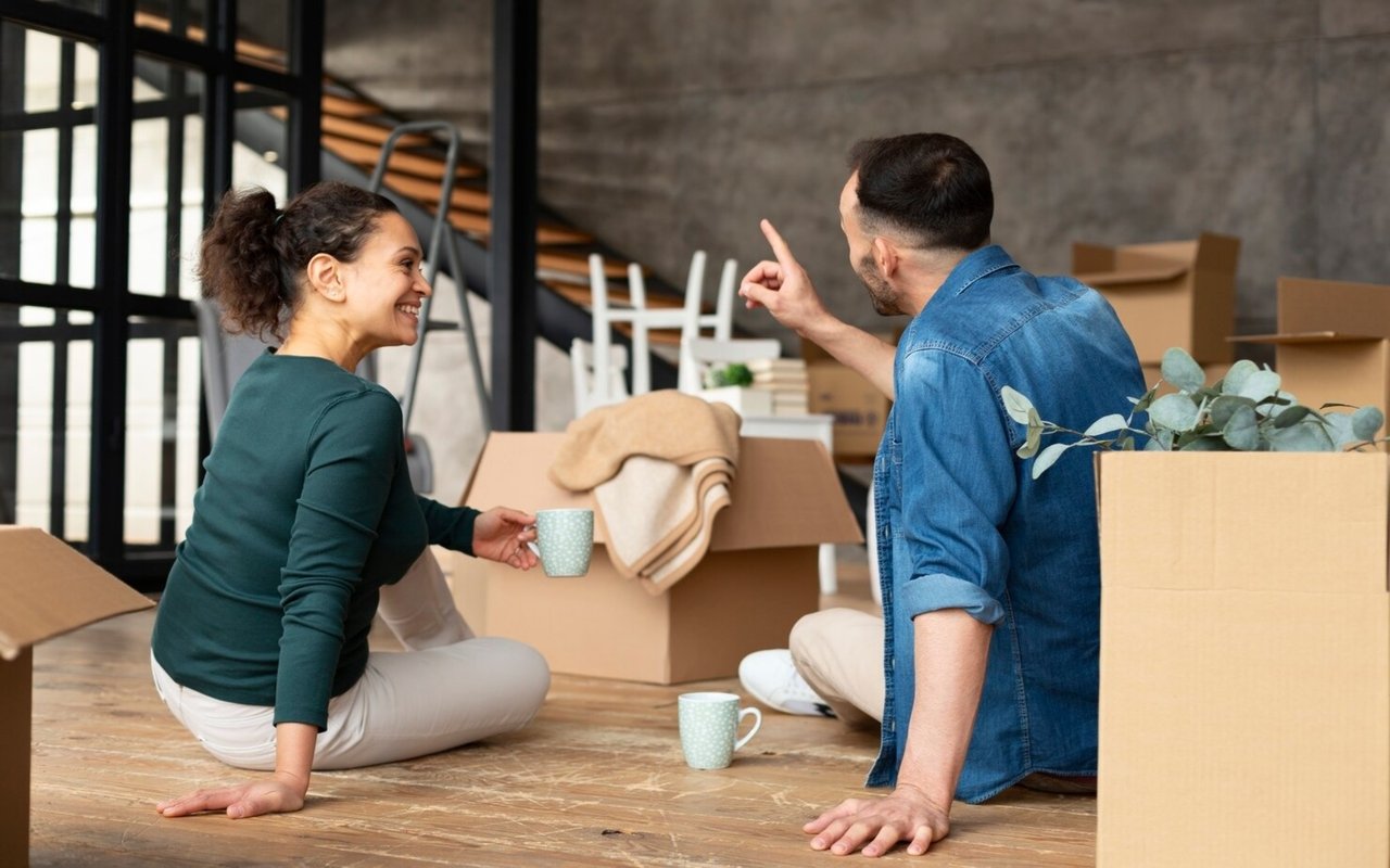 Two people take a break among moving boxes in a partially furnished room.