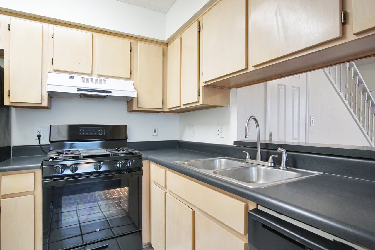 A kitchen with light wood cabinets, black countertops, a double sink, and a black stove with an overhead range hood.