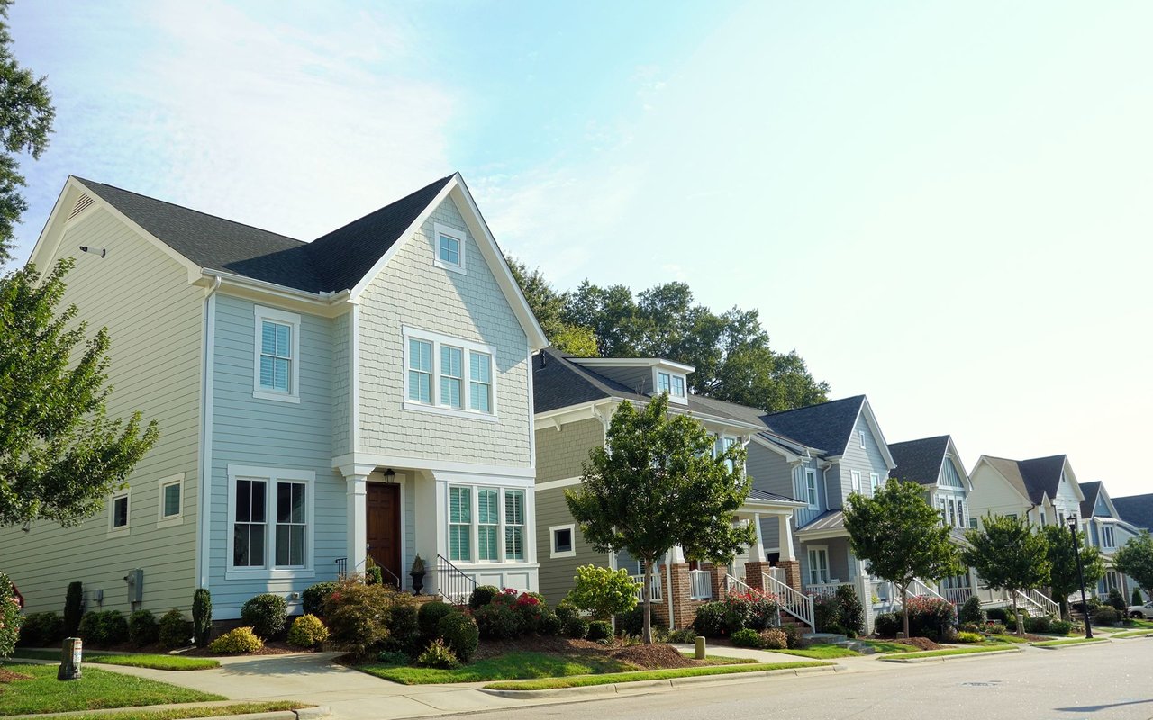 Two-story homes line a landscaped street, with a light blue shingle-sided house in the foreground.