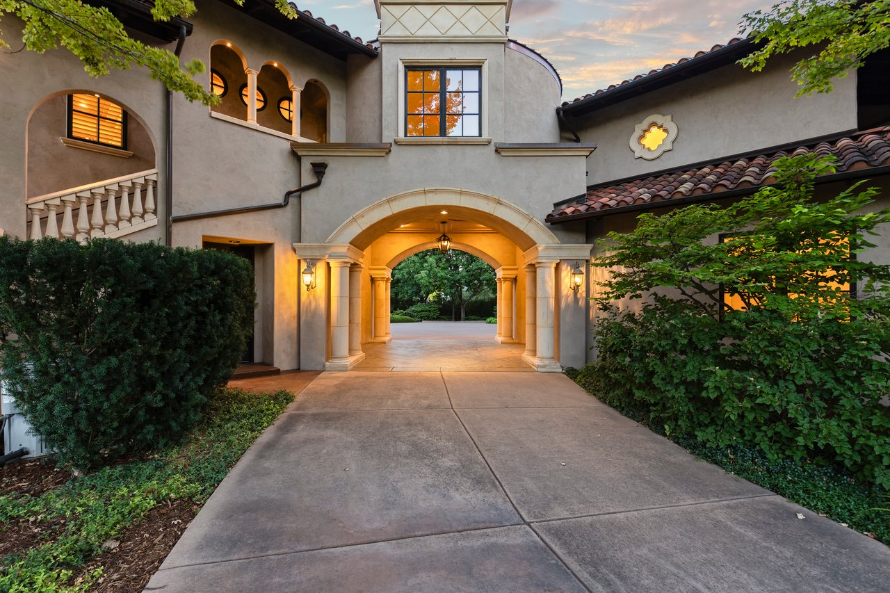 Estate entrance with a central stone archway, broad driveway, and mature garden shrubs.