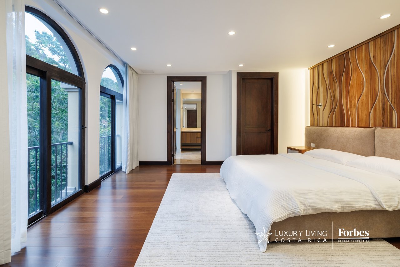 Penthouse bedroom with a white bed, sculptural wood headboard wall, and tall arched windows overlooking trees.