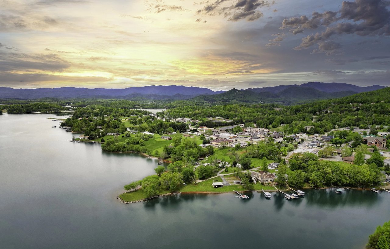 Aerial view of a lakeside town beneath wooded mountains at sunset.