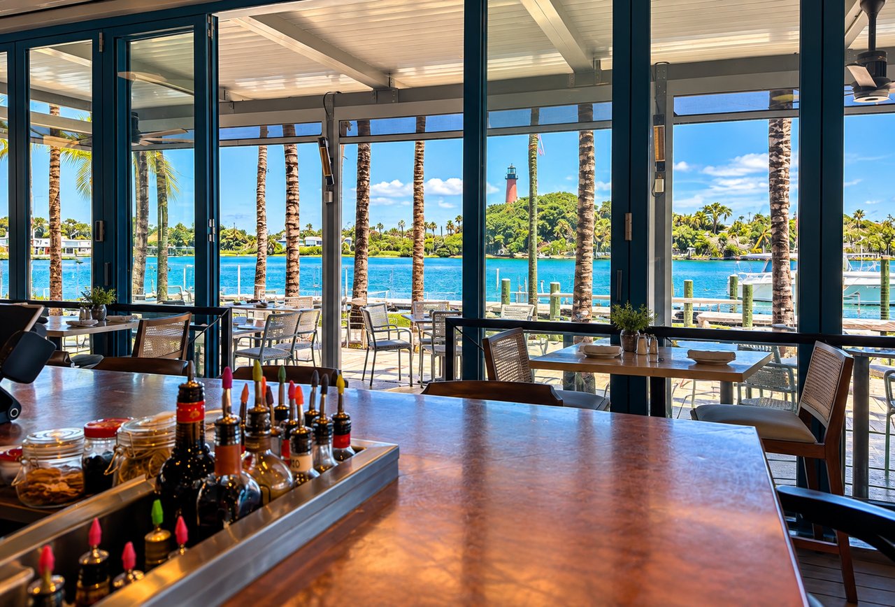 Waterfront restaurant with floor-to-ceiling windows overlooking boats, palm trees, and a lighthouse.