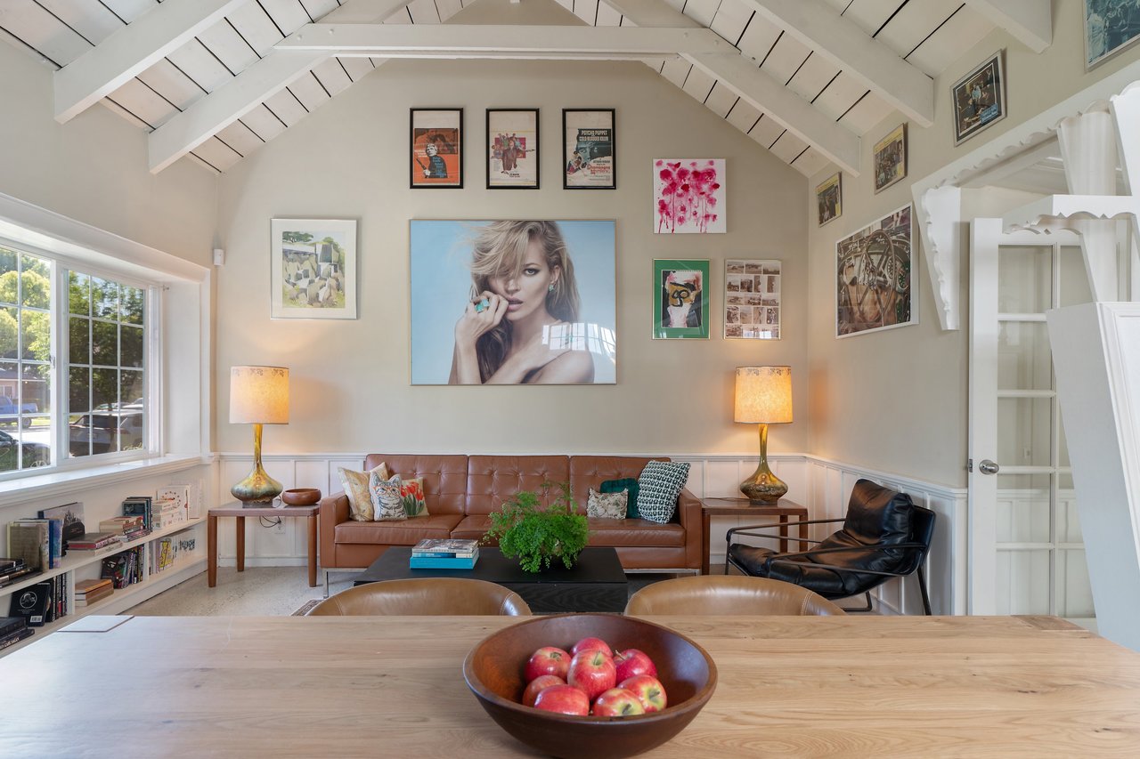 Living room with vaulted ceiling, art-filled wall, brown leather sofa, and large window, viewed from a dining table with apples.
