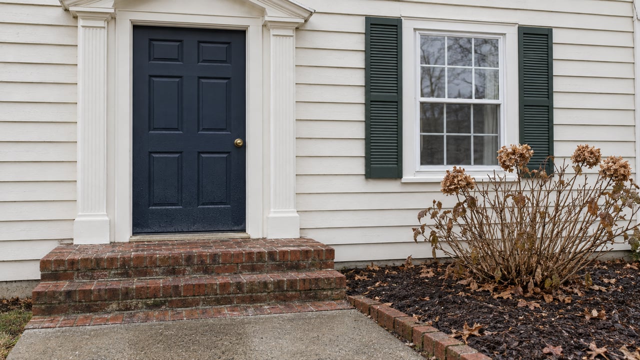 Navy front door and shuttered window on a pale clapboard home, with worn brick steps and dormant oakleaf hydrangea.