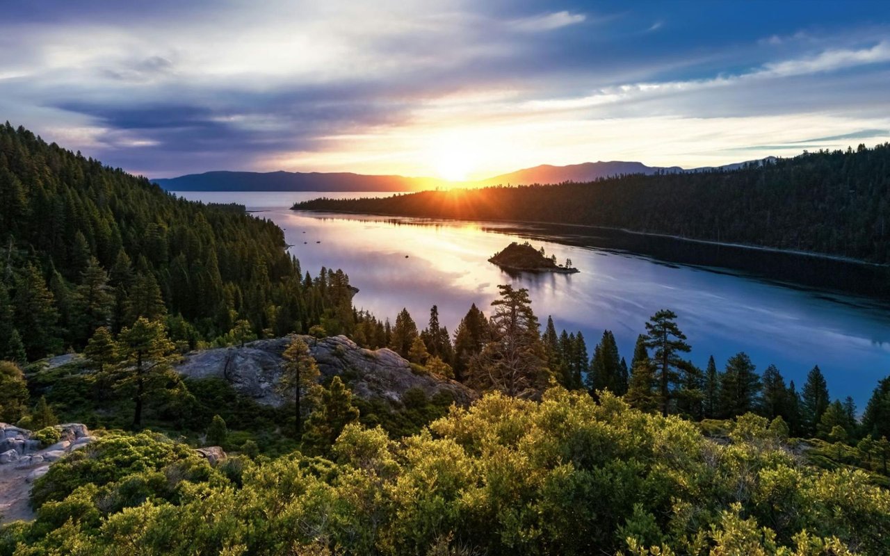  A scenic landscape featuring a wide, calm lake at sunset or sunrise, with a bright sunburst near the horizon reflecting on the water.