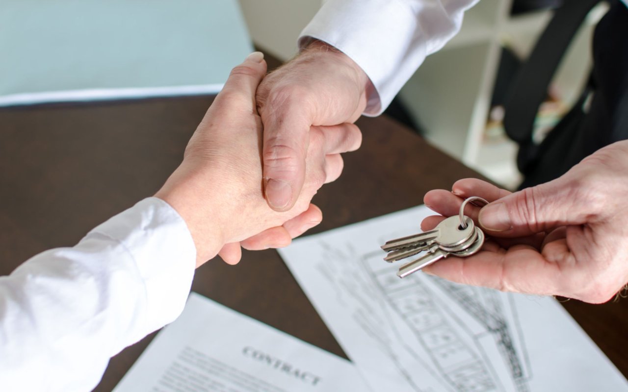 A handshake beside house keys and real estate contract paperwork.