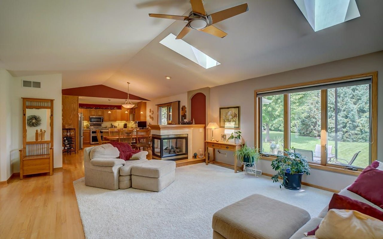 Living room with vaulted skylit ceiling, two-sided fireplace, and windows overlooking a green yard.
