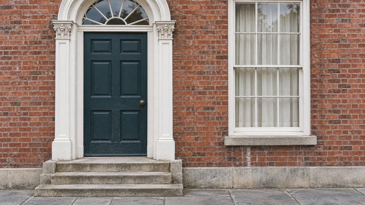 A blue-green timber door with white carved trim and a glazed fanlight beside a sash window in an aged brick townhouse facade.