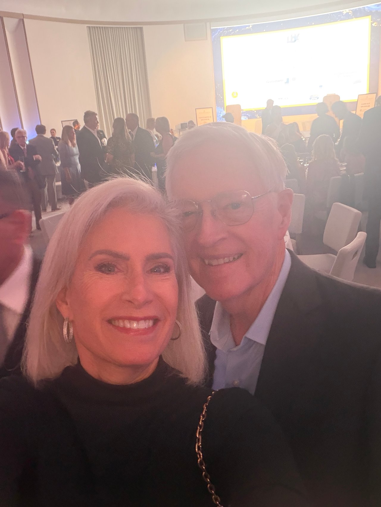 A woman and man smile for a selfie at a crowded awards event.
