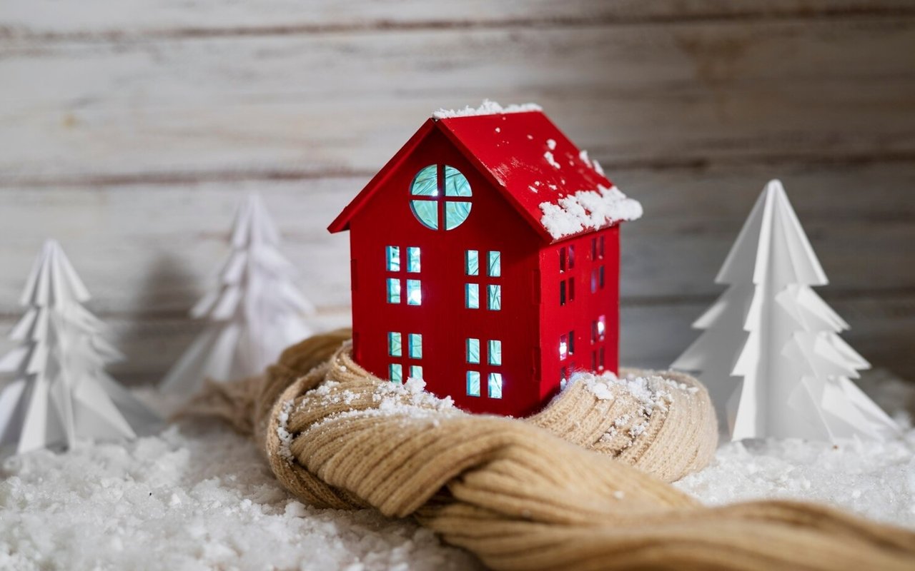 Red model house dusted with snow resting in a knitted mitten among white paper trees.