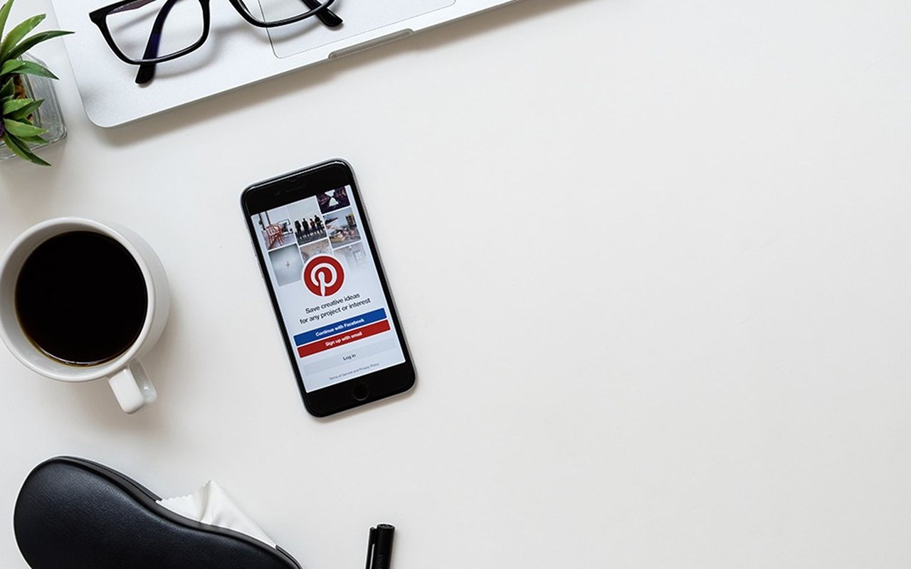 A phone displaying Pinterest beside coffee, eyeglasses, and a small plant on a white desk.