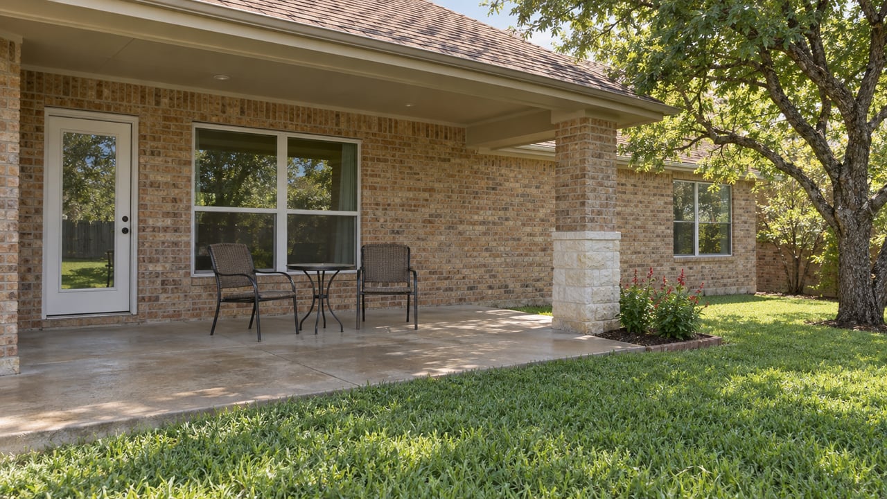 Brick home's covered rear patio with two woven chairs, a concrete terrace and a spreading ebony tree beside the lawn.