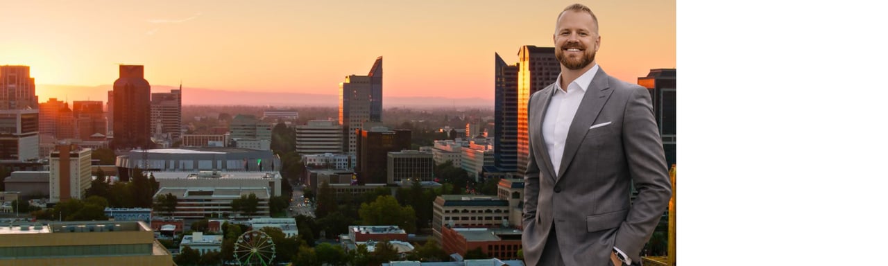 Smiling man in a gray suit with the Sacramento skyline at sunset.