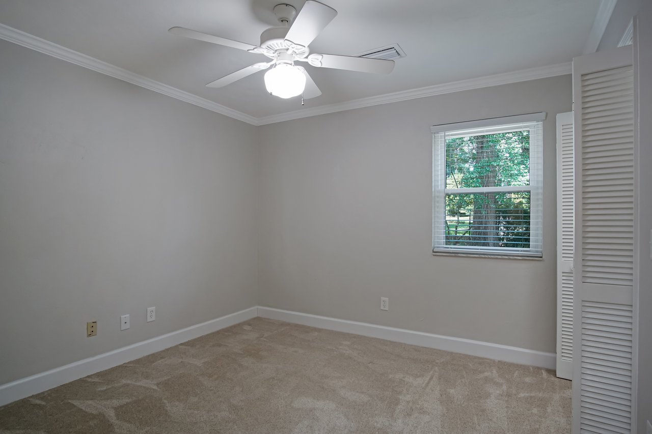 Empty room with beige carpet, light gray walls, and a white ceiling fan. A small window with blinds shows greenery outside, evoking a calm atmosphere.