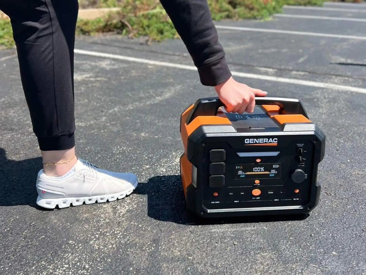 A person carries a black and orange Generac portable power station across pavement.