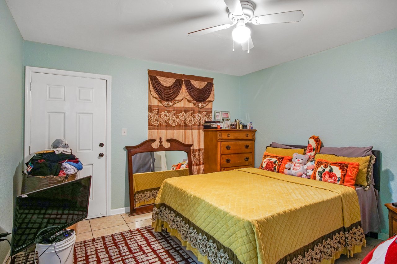Bedroom at 2506 Nugget Lane in Tallahassee, Florida, featuring tile flooring, light green walls, and a ceiling fan with light fixture. The room is furnished with a bed dressed in a yellow coverlet and colorful pillows, a wooden dresser, and a mirror. A decorative curtain covers the window, and a side door provides additional access.