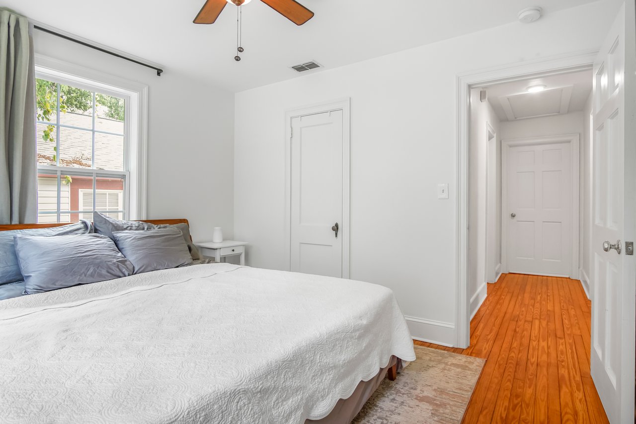 A bright bedroom with white walls features a neatly made bed with gray and blue pillows. A wooden floor leads to a hallway, creating a calm, inviting atmosphere.