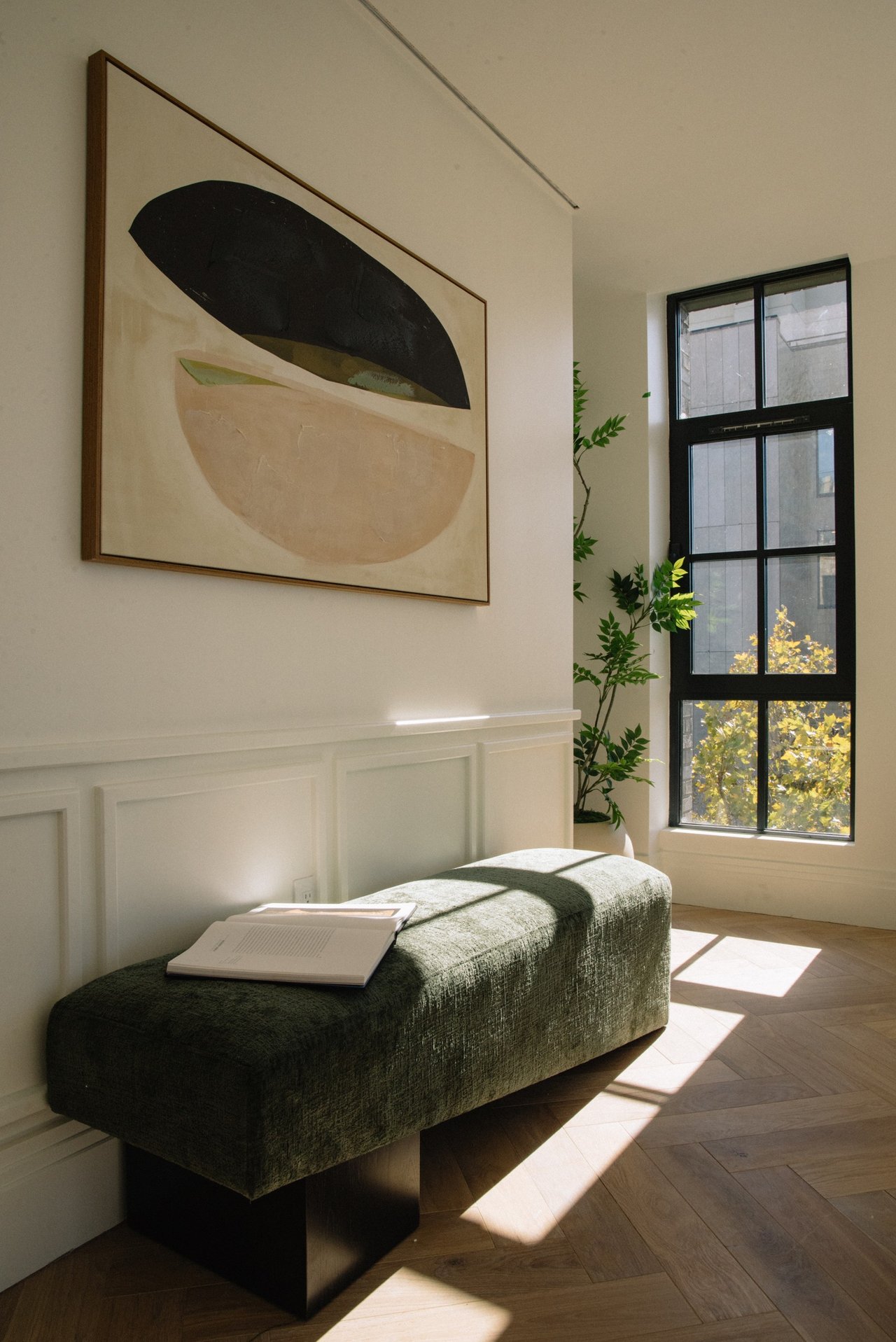 Sunlit sitting area with a green upholstered bench, abstract painting, and tall black-framed window.