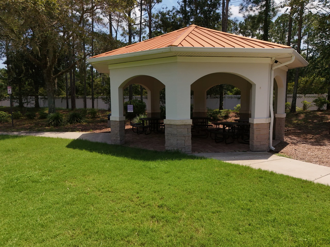 Community pavilion with a terracotta-colored roof, stone columns, and picnic tables beside a green lawn.