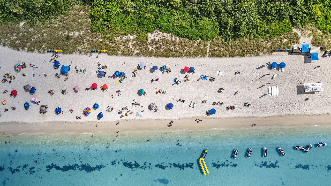 Aerial view of a busy beach with colorful umbrellas and boats in clear turquoise water.