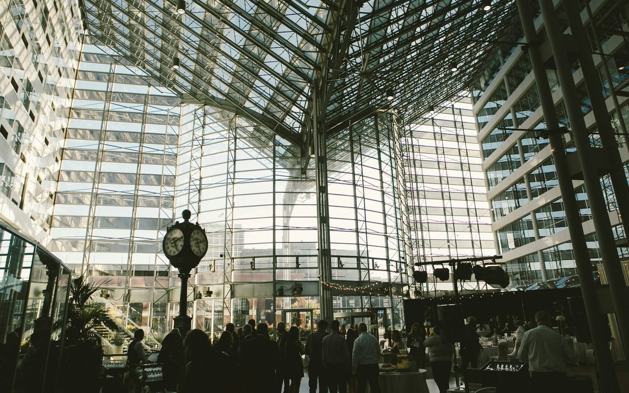 Glass-roofed commercial atrium with steel trusses, a freestanding clock, and an event crowd.