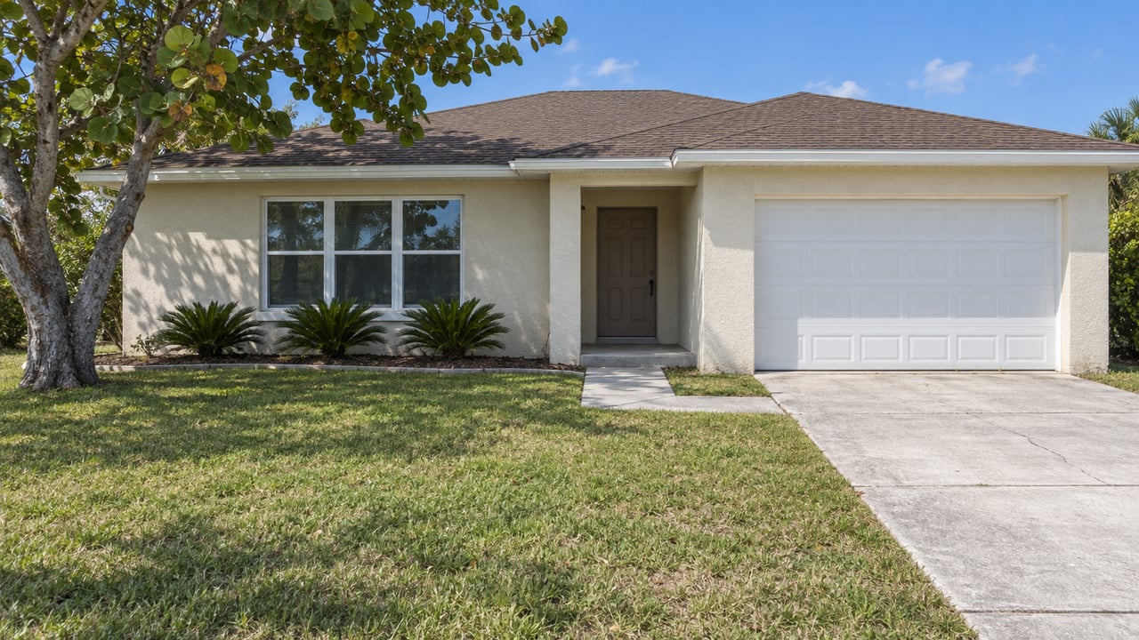 Sunlit single-story stucco home with a brown hip roof, deep lawn, concrete driveway and sea grape tree.