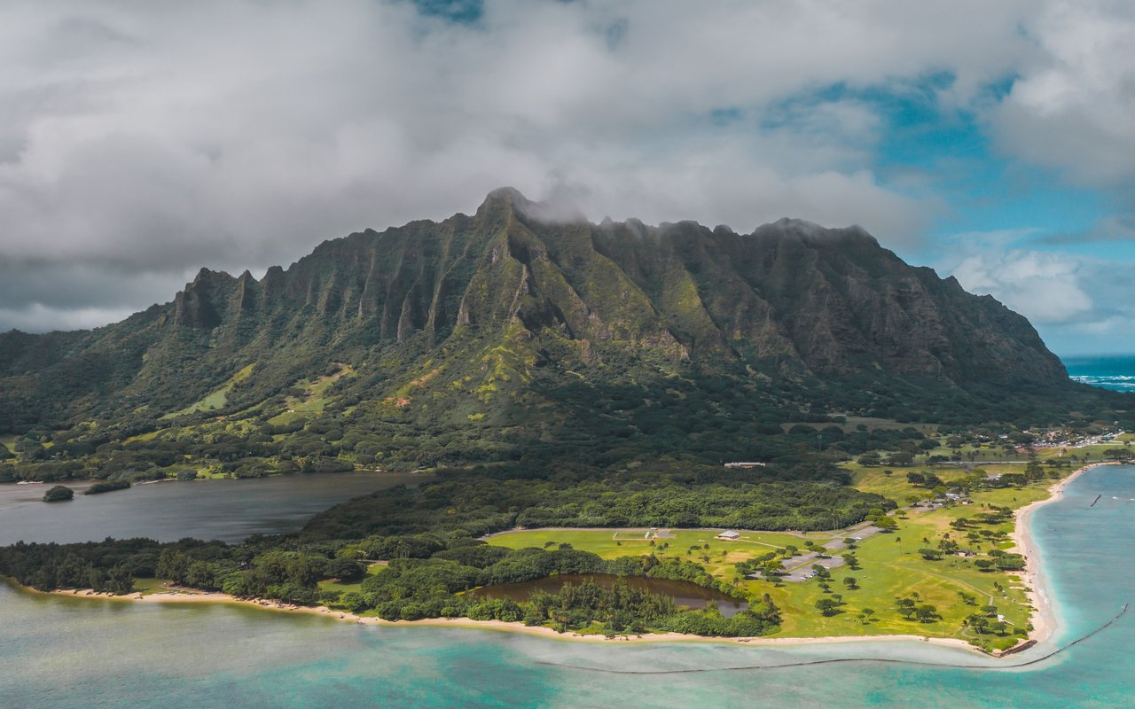Aerial view of a ridged green mountain behind a peninsula with turquoise water and sandy shores.