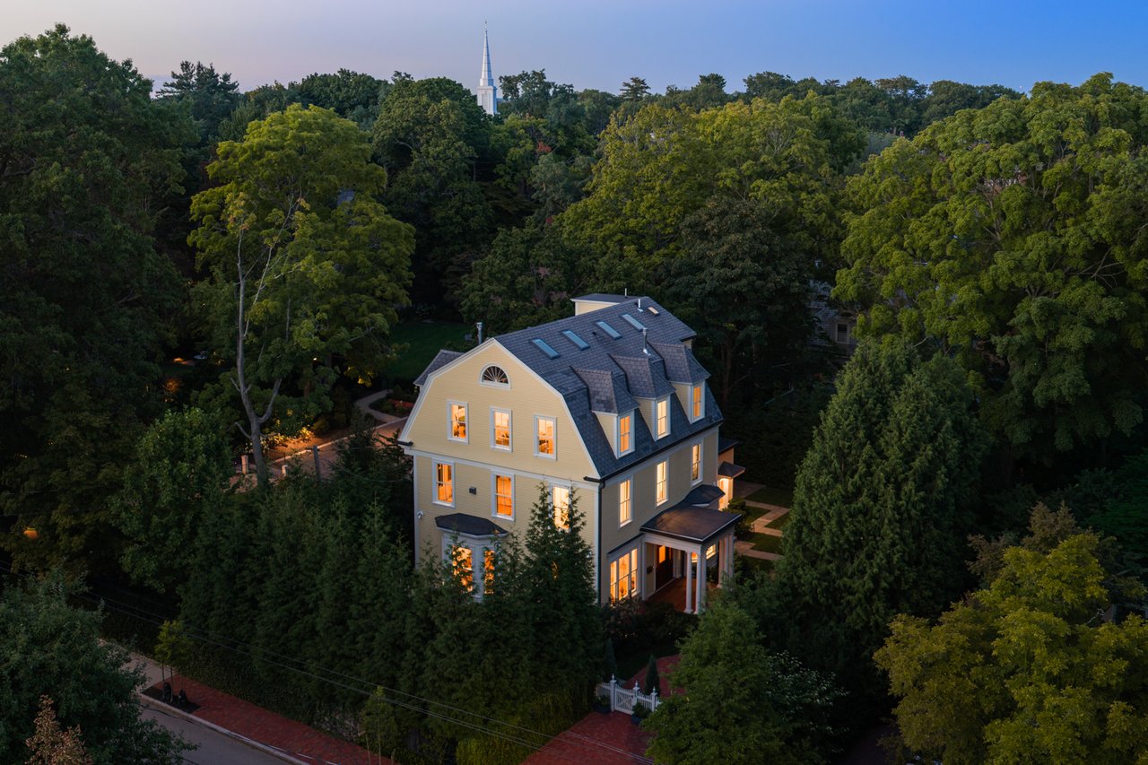 Aerial view of a yellow Victorian house with glowing windows, steep roof, and wooded grounds at dusk.
