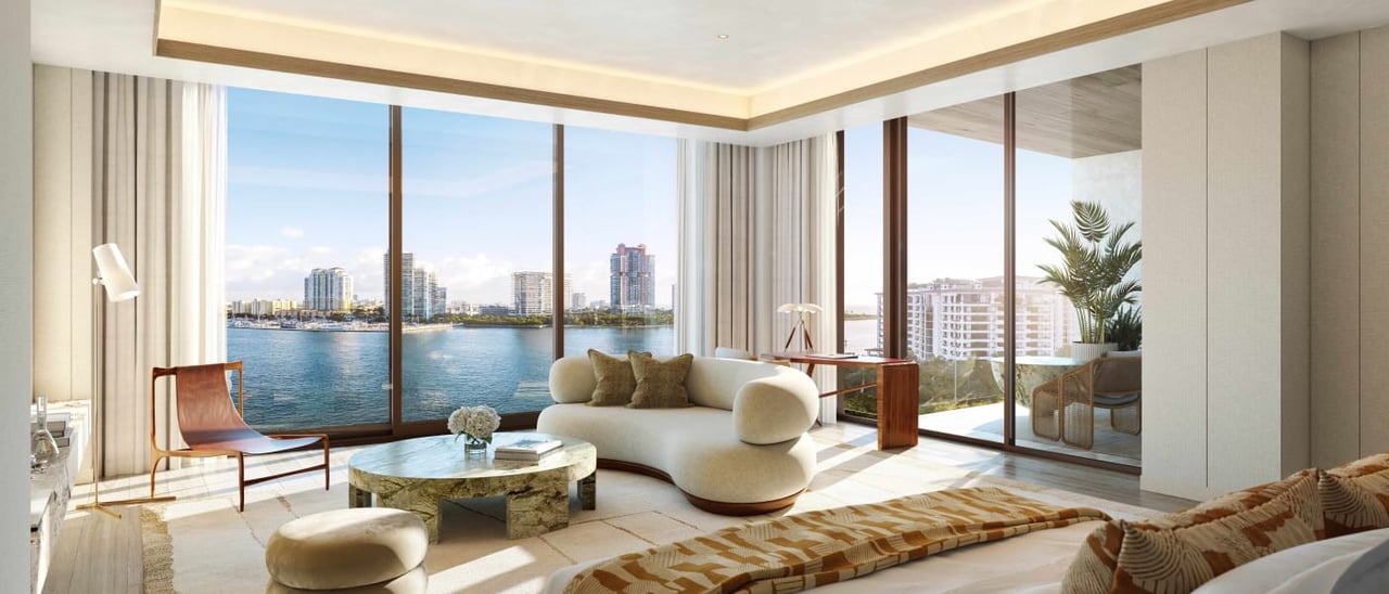 Furnished living room with floor-to-ceiling windows overlooking water and a distant city skyline.