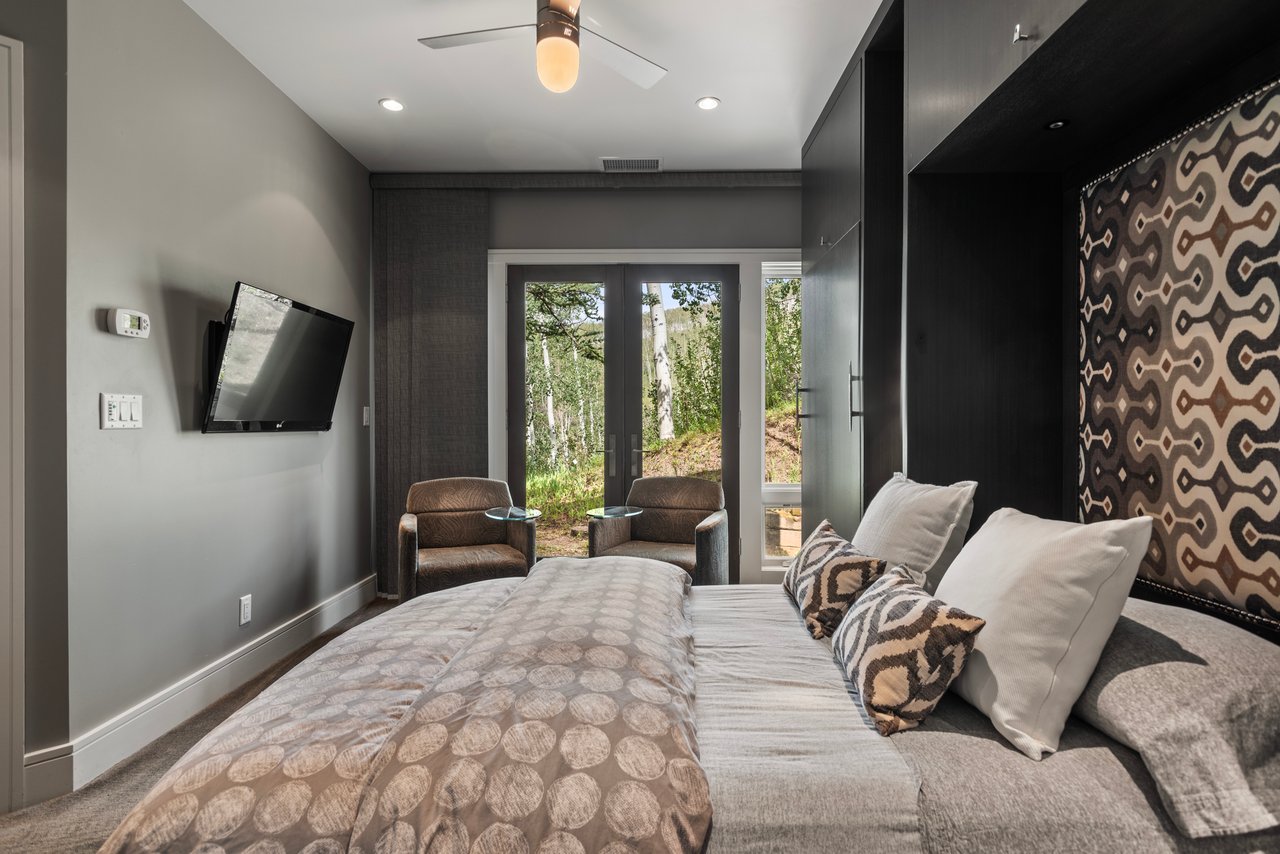 Bedroom with a large bed, patterned accent wall, and glass doors overlooking trees.