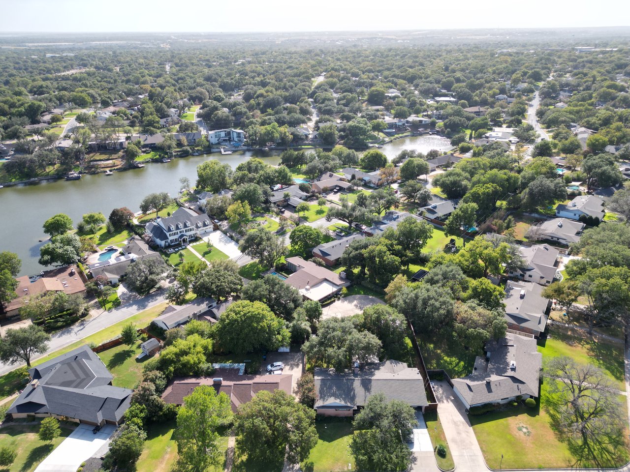 Aerial view of homes and tree-lined streets surrounding a lake, with a white house and pool near the shore.