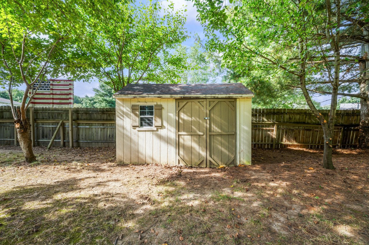 Wooden backyard shed with double doors and a small window, shaded by mature trees inside a fenced yard.