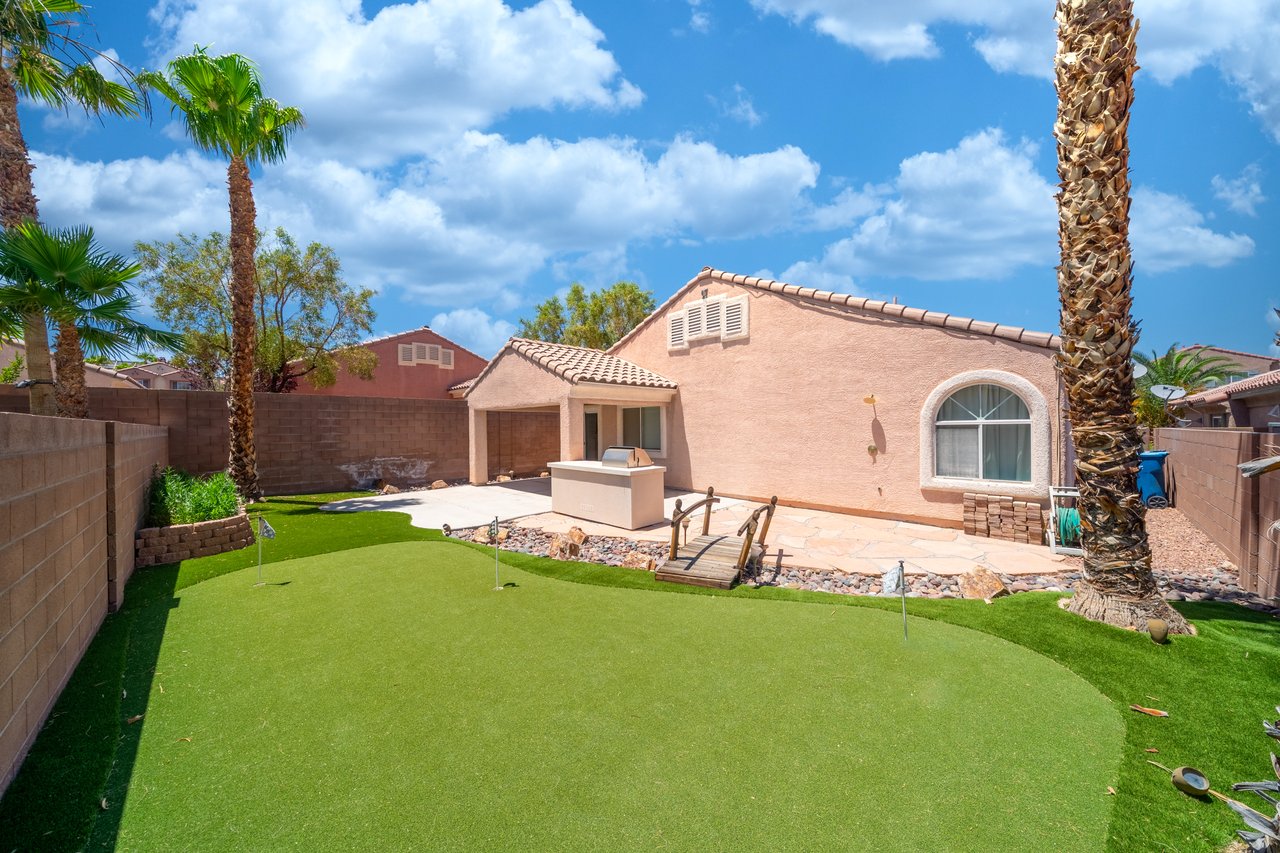 Home backyard with artificial turf putting green, palm trees, and a covered patio with a grill.