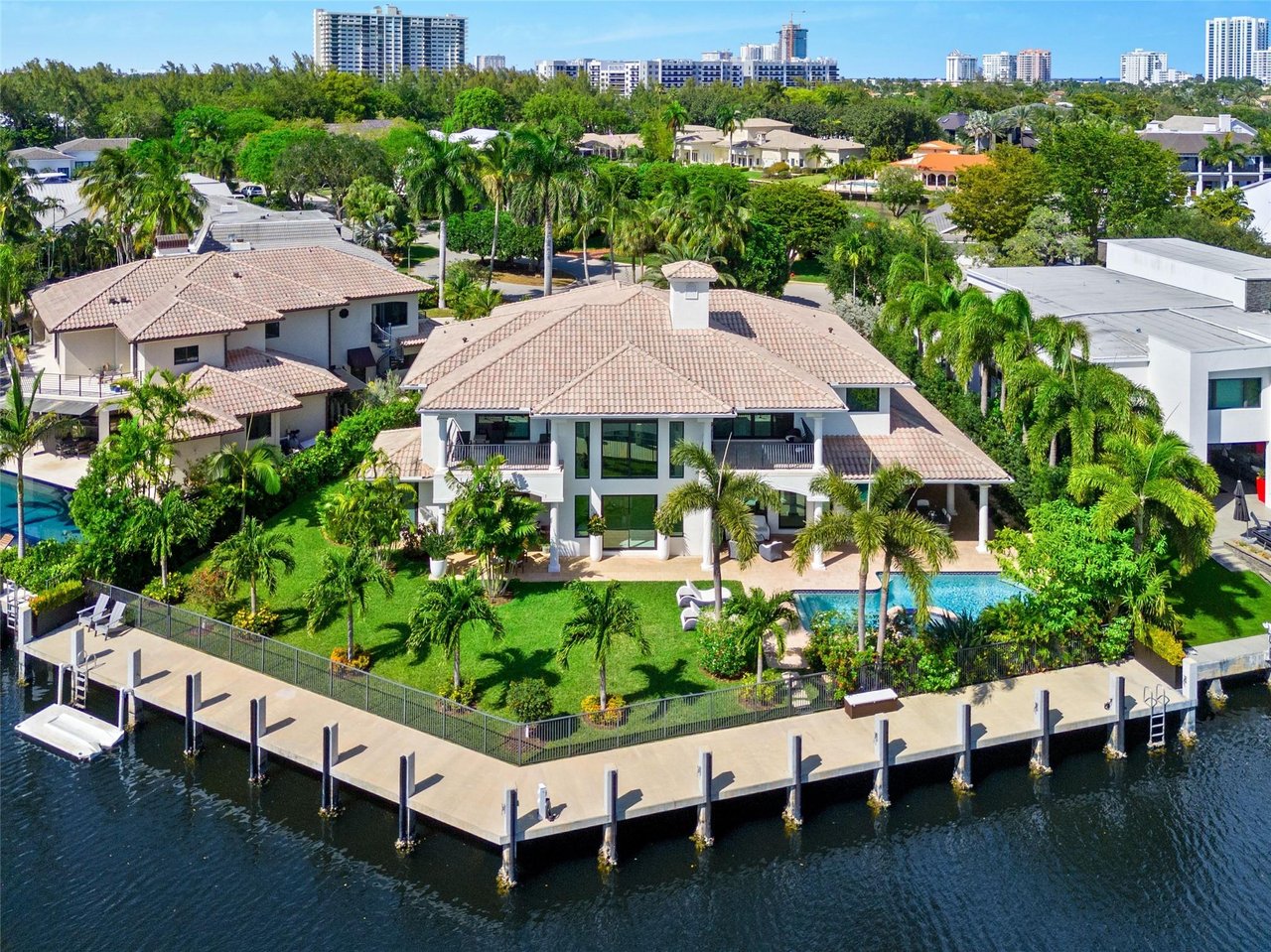 Waterfront home with a swimming pool and long canal-side dock, surrounded by palms and neighboring homes.