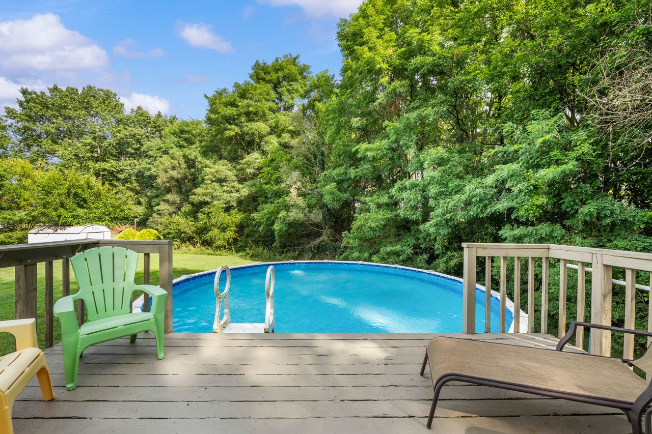 Above-ground pool beside a wooden deck with green chairs and a wooded backyard.