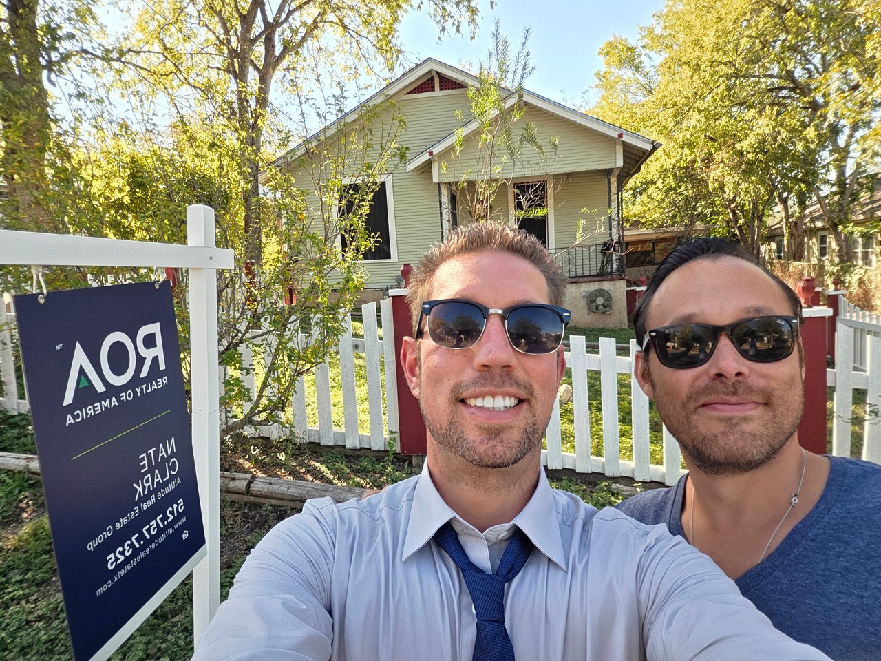 Two men pose for a selfie in front of a pale green house and real estate sign.