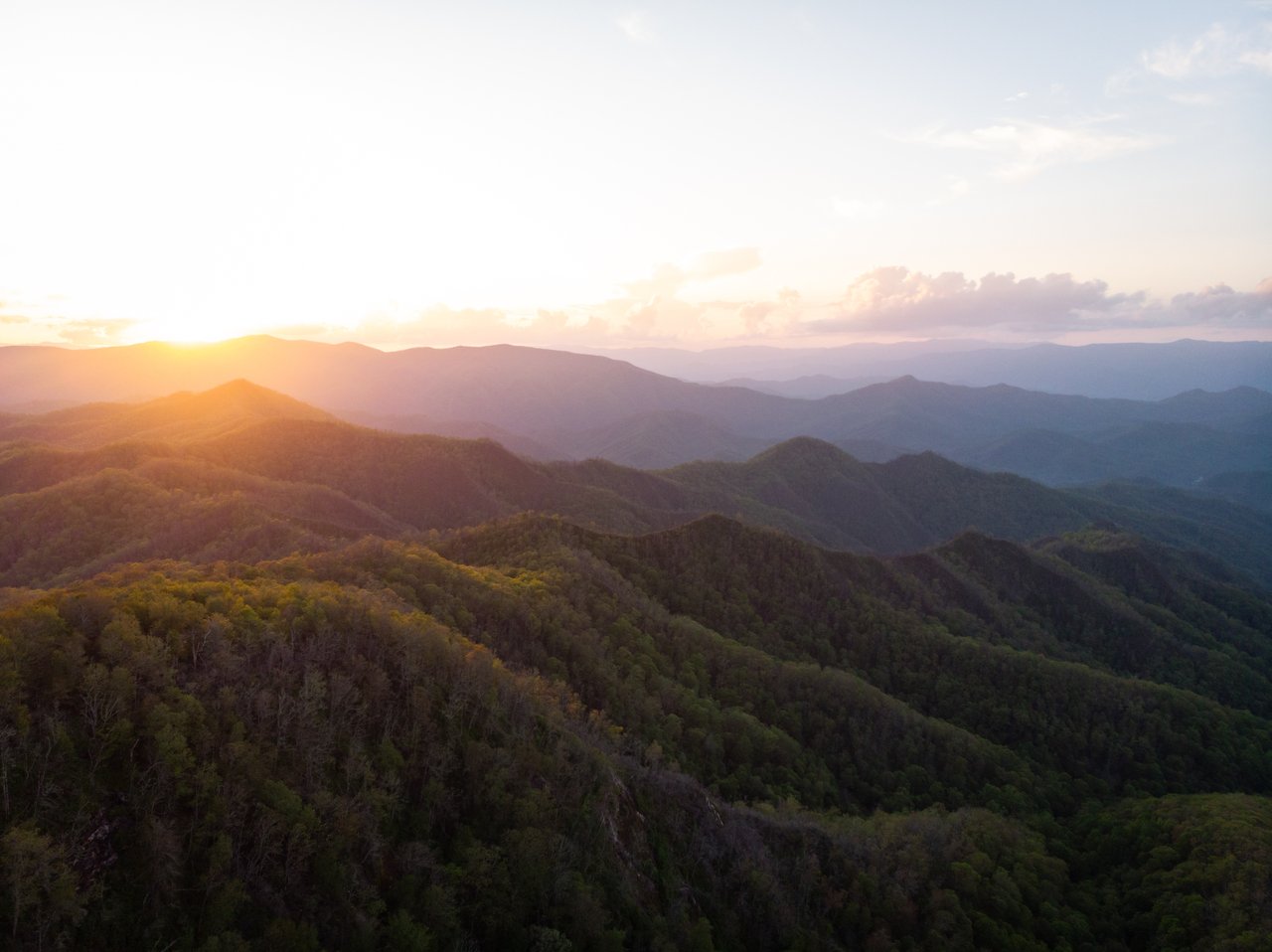 The sun sets over rolling forested mountains, painting a serene backdrop of clear skies and soft clouds—a perfect platform for real estate agents to showcase nature’s beauty.