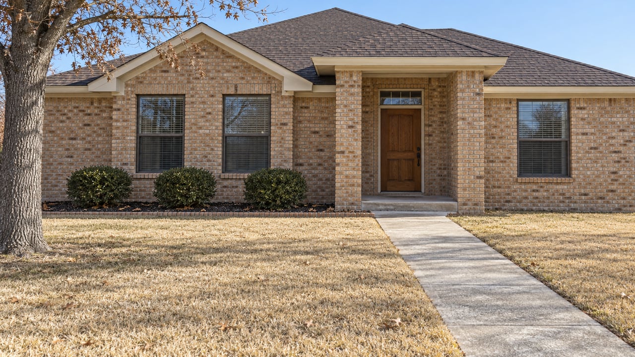 A buff-brick single-story home behind a broad dormant lawn, with a recessed oak entry and carefully edged planting beds.