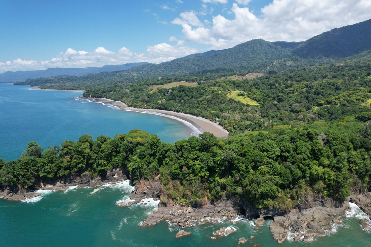 Aerial view of a curved beach and turquoise Pacific water bordered by forested hills and rocky cliffs.