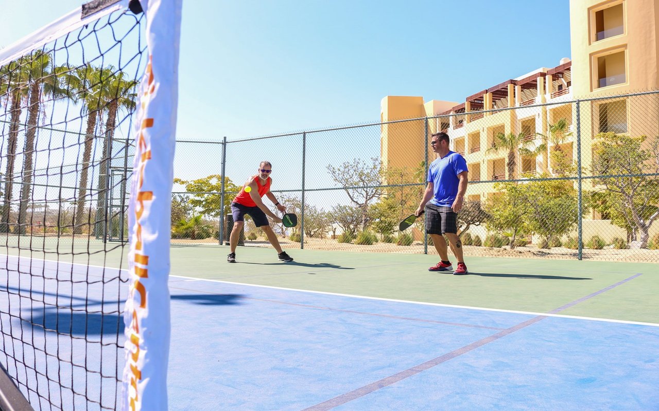 Two men play pickleball on a blue-and-green outdoor court beside a yellow residential building.