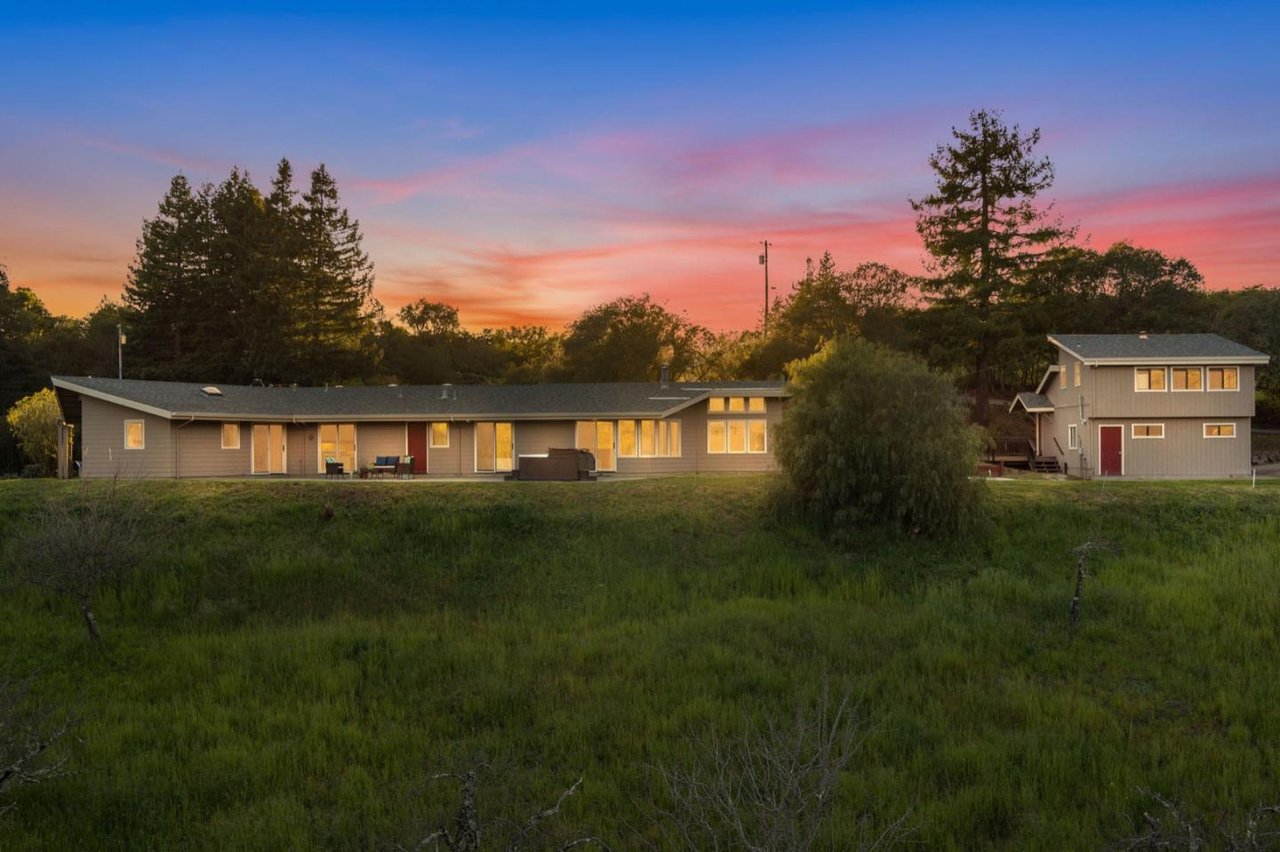 Long single-story home and two-story outbuilding beneath a colorful sunset sky.