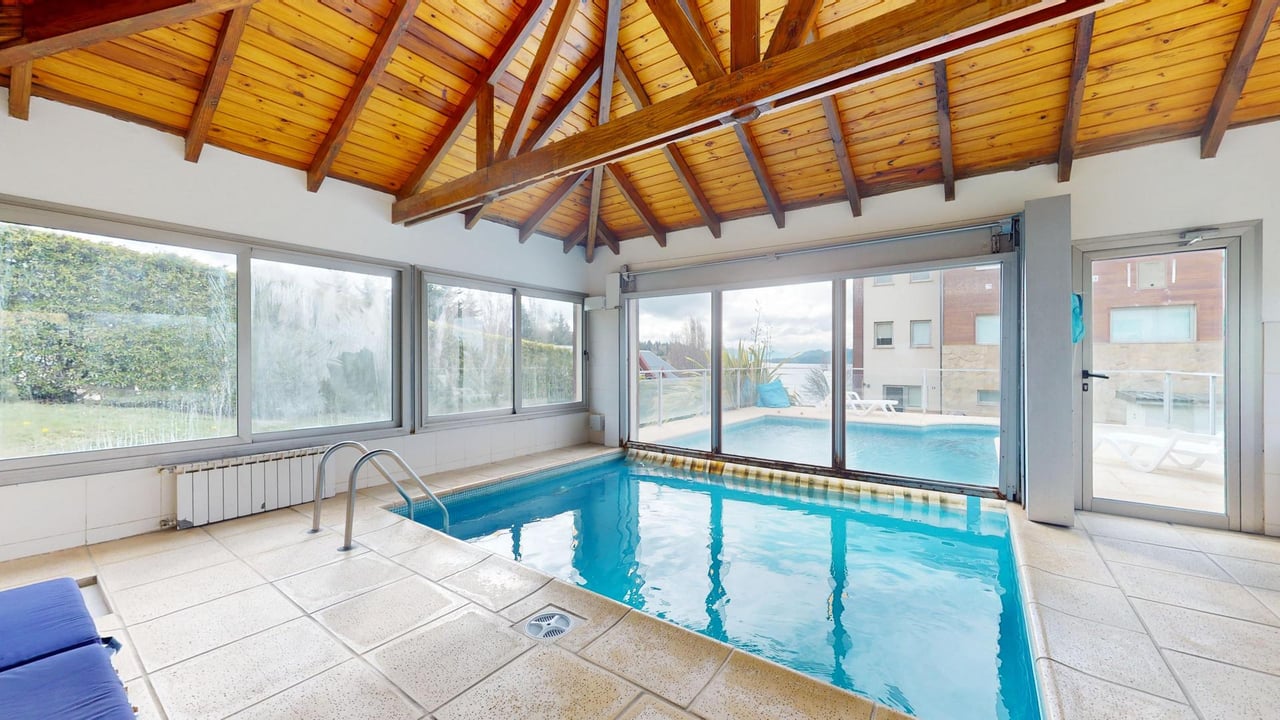 Indoor swimming pool beneath a vaulted wooden ceiling, with glass doors opening toward an outdoor pool.