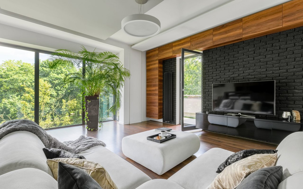 Modern living room with a white sectional, black brick media wall, and large windows overlooking greenery.