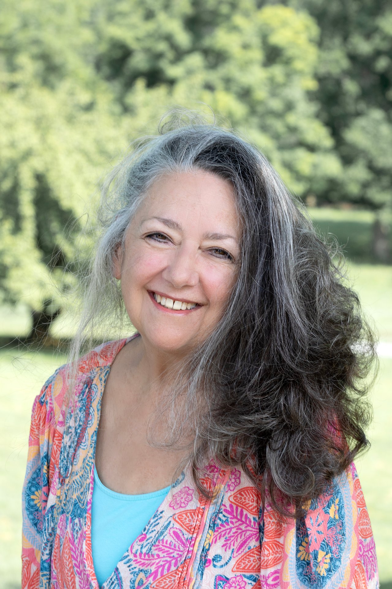 Debra Gurin smiling outdoors, with long gray hair and a colorful patterned cardigan.