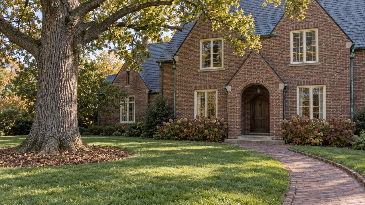 A mature white oak stands across a broad lawn from a brick house with a slate roof and arched entry.